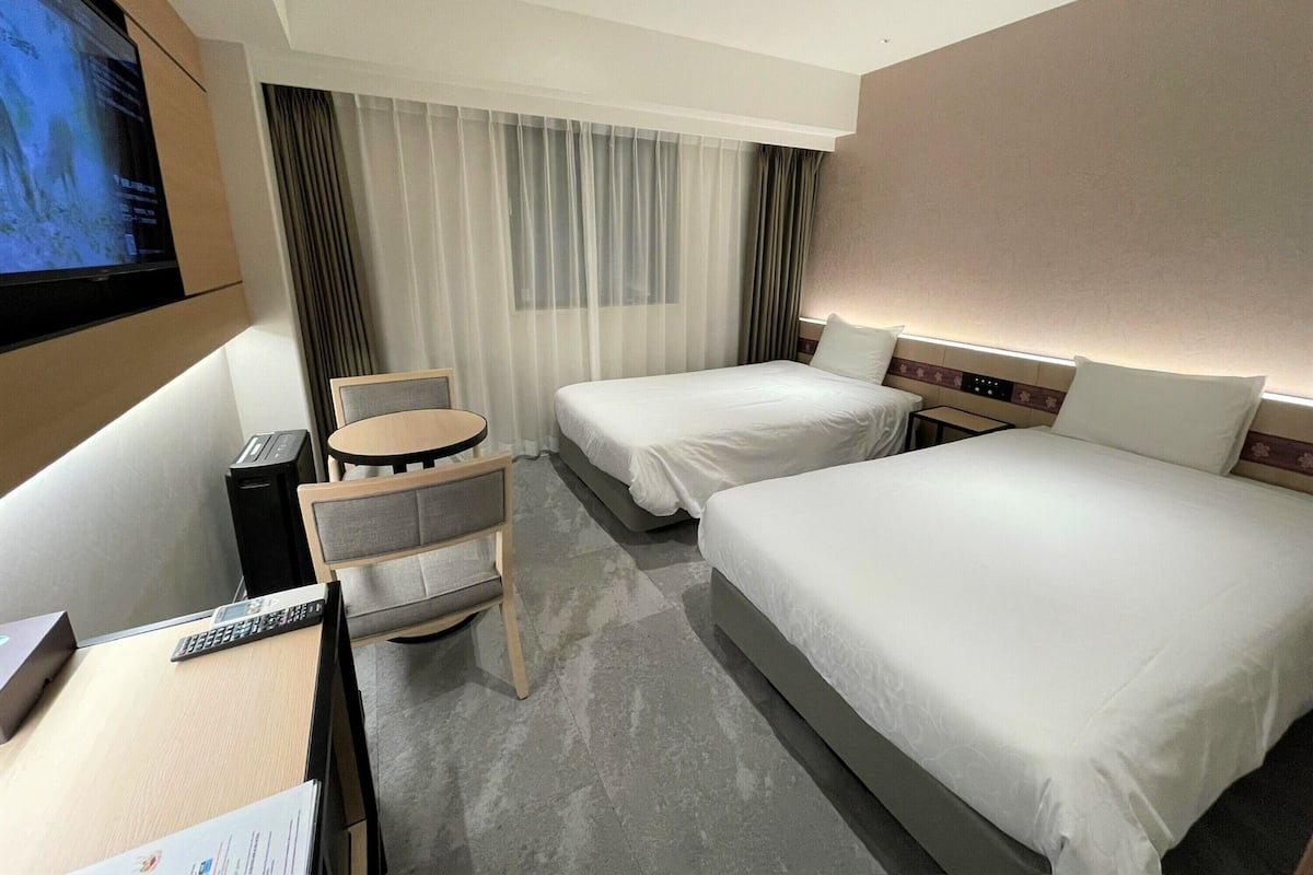 A hotel room with two beds, a desk, a chair, a TV, and a window with curtains.