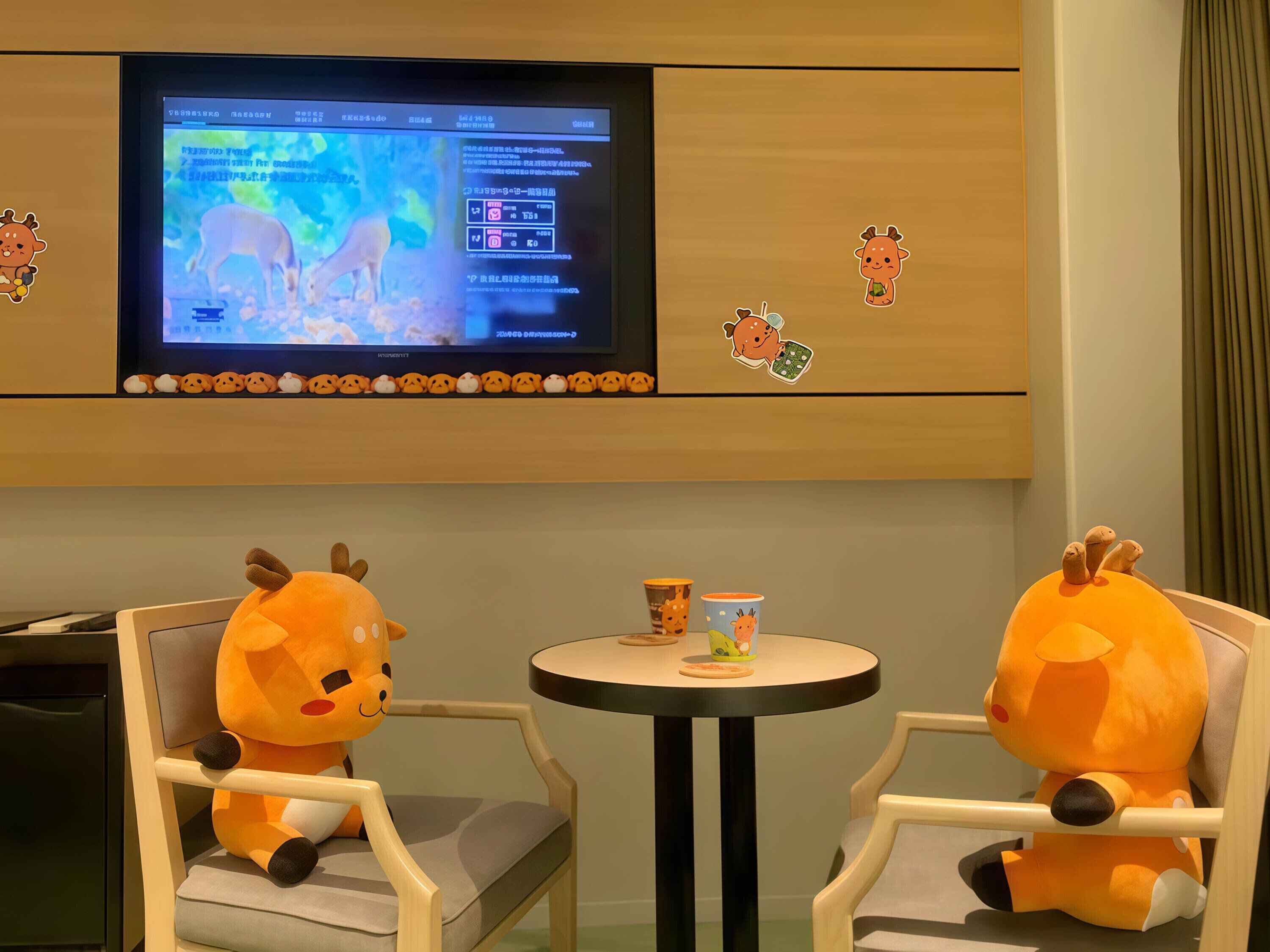 Una habitación con un televisor montado en la pared, dos peluches en las sillas, una mesita con una taza y una caja, y una fila de pegatinas de animales en la pared.