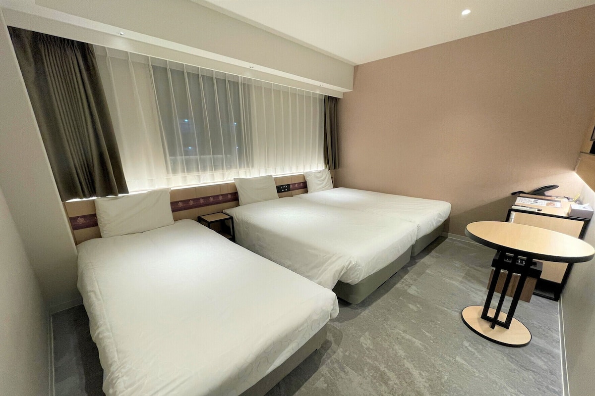 A hotel room with two beds, a small table, and a window with curtains.