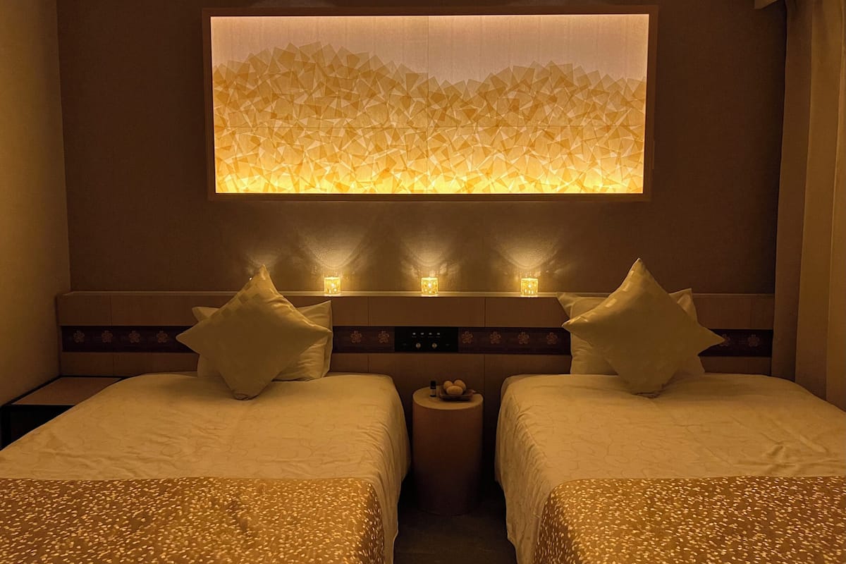 A hotel room with two beds, a nightstand, and a wall-mounted light fixture.