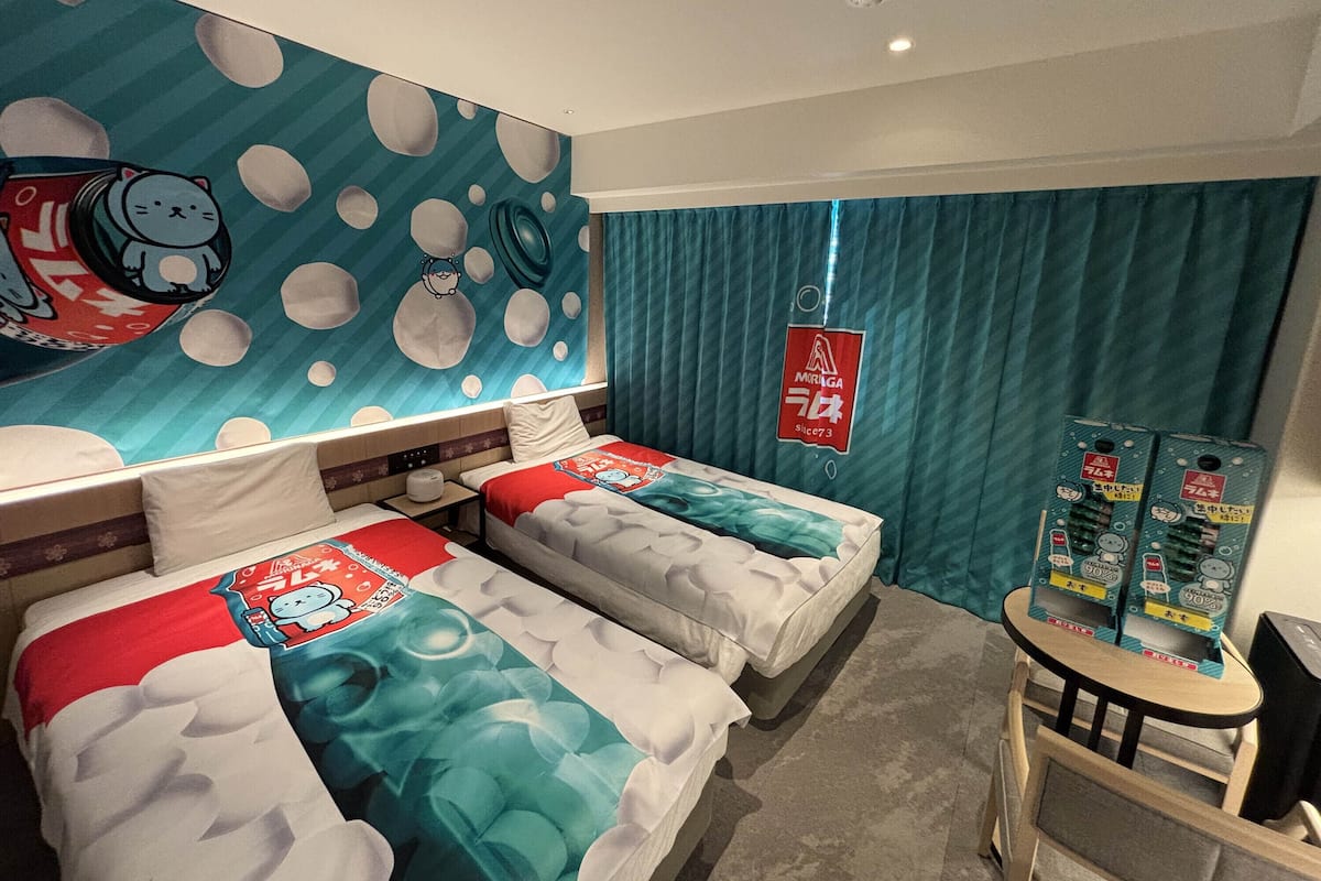 A hotel room with two beds, a mural on the wall, a small table, and a curtain.