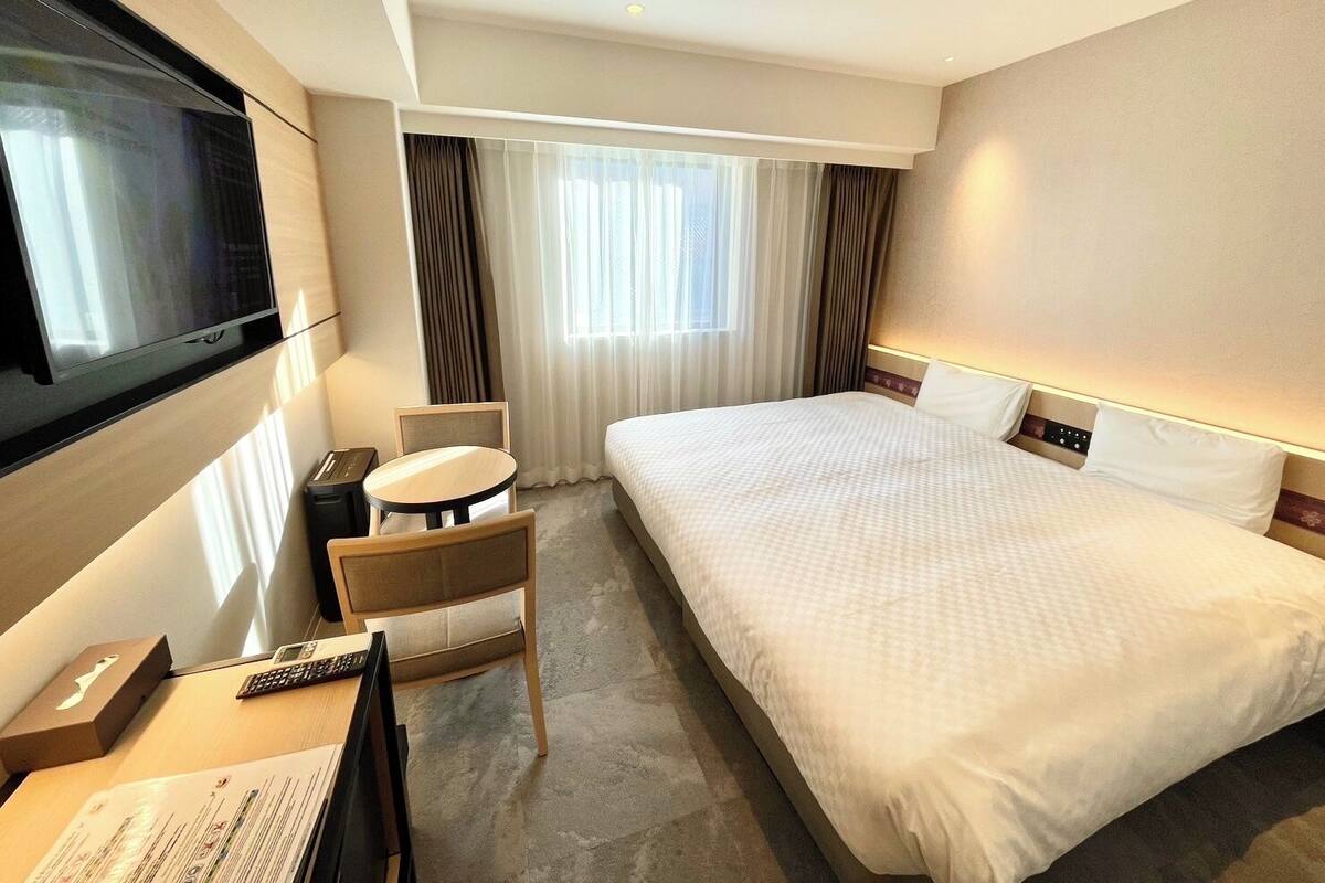 A hotel room with a large bed, a desk with a chair, a TV mounted on the wall, and a window with curtains.