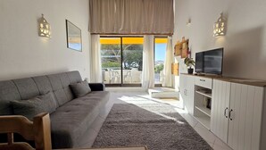 Een woonkamer met een grijze bank, een televisie op een houten kast, en uitzicht op een balkon met tuinmeubilair.