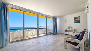 Un balcon spacieux offrant une vue dégagée sur la mer et les bâtiments, avec des meubles en osier blancs et un meuble blanc orné d’un vase de fleurs.