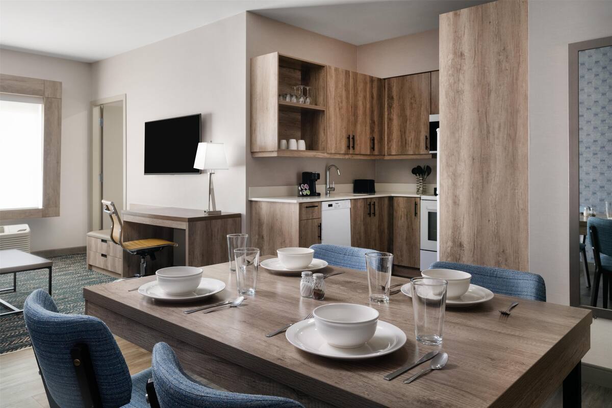 A modern dining area with a wooden table set for four, a television mounted on the wall, and a kitchen area with wooden cabinets.