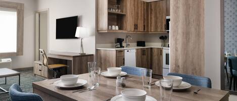 Uma sala de jantar moderna com uma mesa de madeira posta para quatro pessoas, uma televisão na parede e uma área de cozinha com armários de madeira.