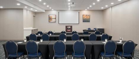 A conference room with a long table, blue chairs, a projector screen, and framed pictures on the wall.