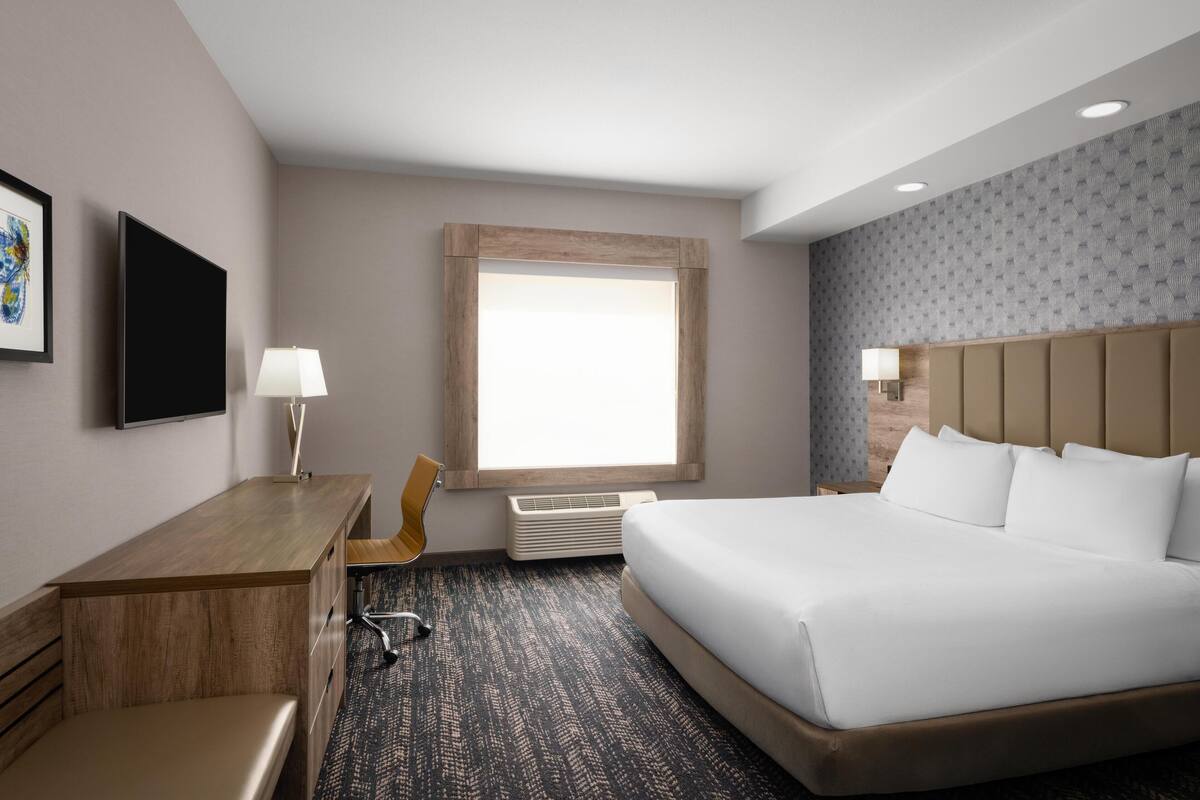 A hotel room with a large bed, a desk, a chair, a flat-screen TV, and a window with natural light.