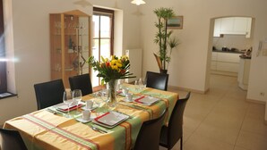 Un coin repas avec une table dressée pour quatre personnes, des chaises, un vase avec des fleurs et, au fond, une cuisine visible.