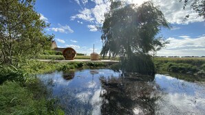 Un paysage paisible avec un étang, une structure en bois qui ressemble à un sauna et un grand arbre dont le reflet se mire dans l’eau.