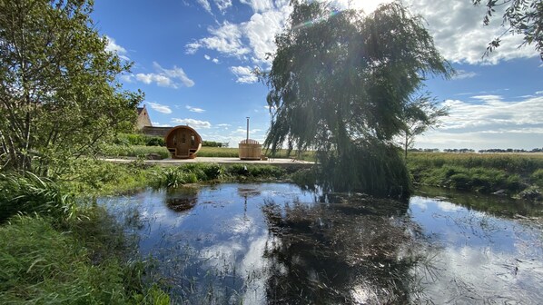 A serene landscape with a pond, a wooden structure resembling a sauna, and a large tree reflecting in the water.