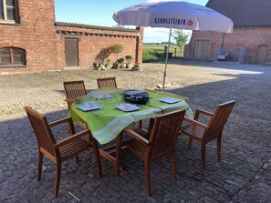 Un área de comedor al aire libre con una mesa dispuesta para cuatro personas, rodeada de sillas de madera, bajo un toldo con el logo de la marca.