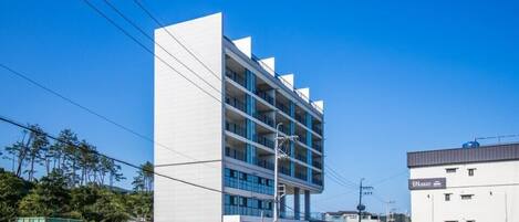 A modern multi-story hotel with a white facade and blue accents, featuring balconies and a parking area.
