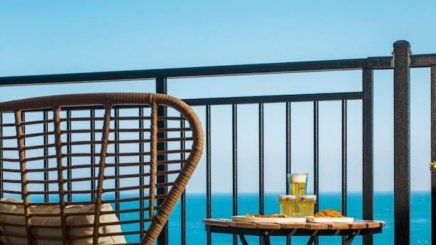 Una silla de mimbre y una pequeña mesa con bebidas y aperitivos en un balcón con vista al océano.