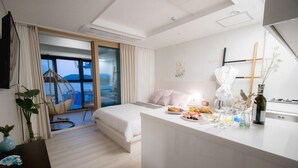 Habitación de hotel moderna con cama, zona de comedor con comida y bebidas, vista al mar y balcón con una silla.