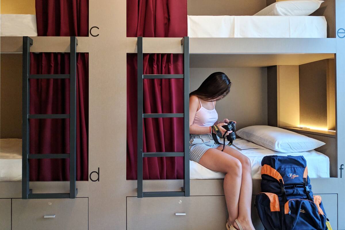A bunk bed room with a person sitting on the lower bunk, holding a camera. There are two ladders, one on each side of the bed, and a suitcase on the floor.