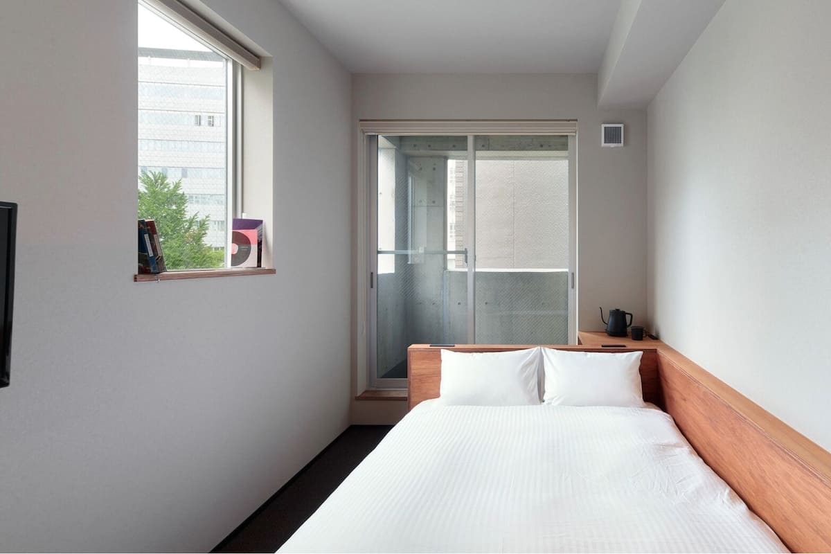 A hotel room with a bed, a window, a door leading to a balcony, and a small table with a kettle and cup.