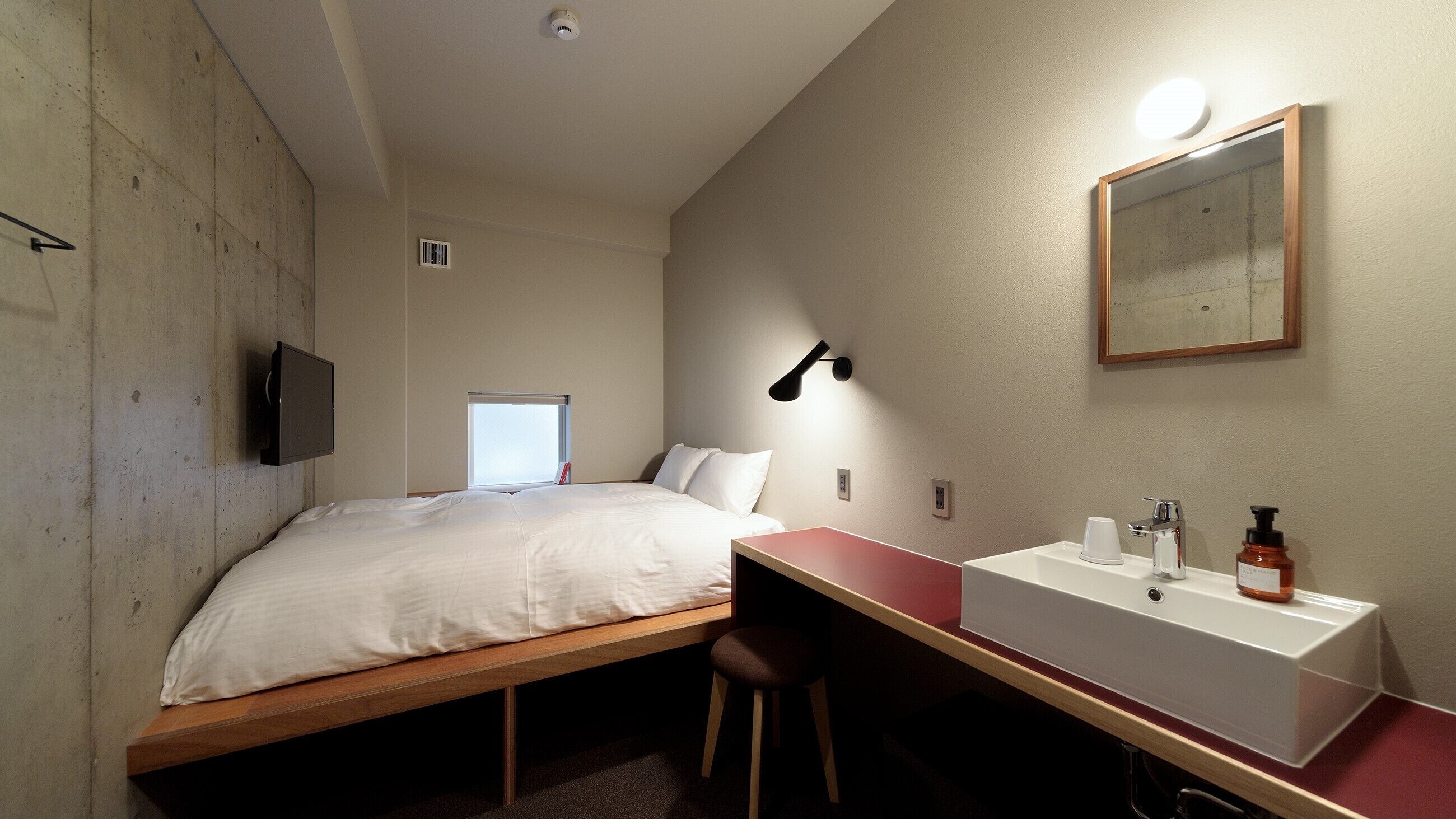 A small hotel room with a bed, a desk, a chair, a sink, and a mirror.