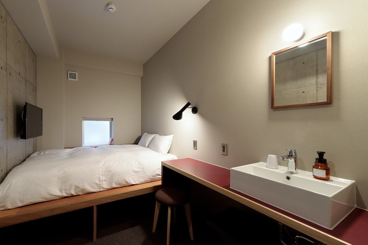 A small hotel room with a bed, a desk, a chair, a sink, and a mirror.