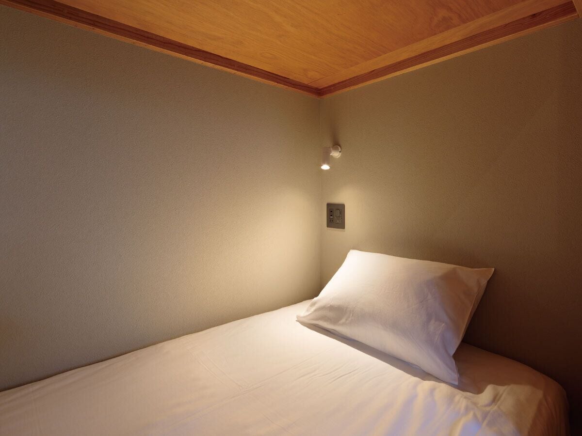 Een eenpersoonsbed met witte lakens in een kamer met houten balkenplafond en een lichtschakelaar aan de muur.