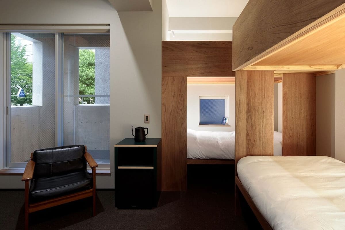 A compact bedroom with a single bed, a chair, a small table with a kettle, and a window providing a view of greenery outside.