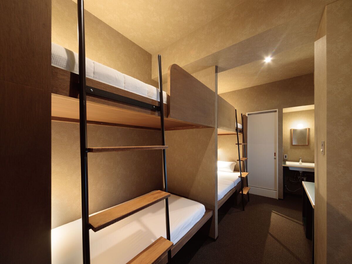 A hotel room with bunk beds, a small desk, a chair, and a bathroom with a sink and mirror.