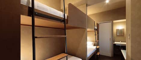 A hotel room with bunk beds, a small desk, a chair, and a bathroom with a sink and mirror.