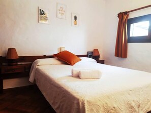 A neatly made bed with a white comforter and an orange pillow, a wooden nightstand with a lamp, and a wall with framed artwork including a pineapple and a leaf.