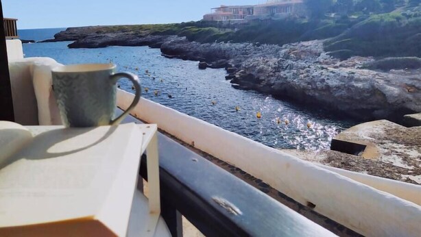 A coastal view from a balcony with a stone railing, a mug on a table, and a building with a rocky shoreline in the background.