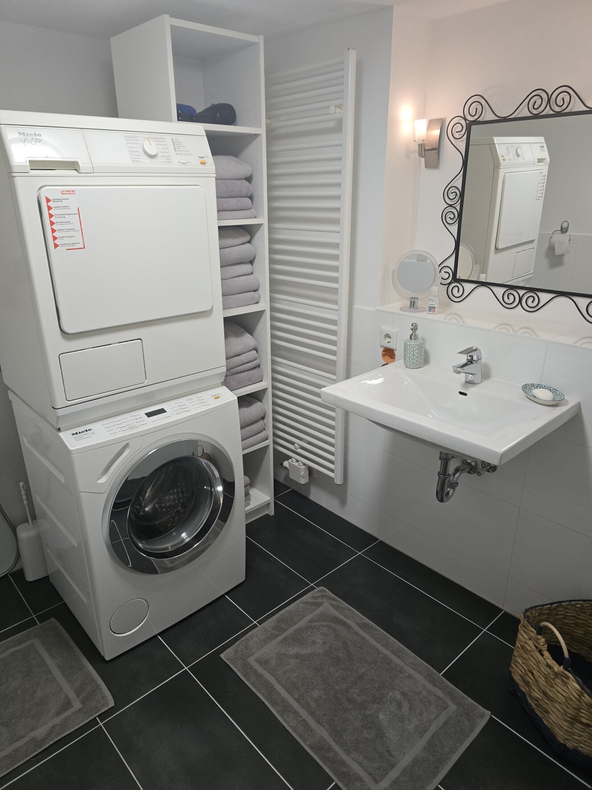 A compact laundry area with a front-loading washing machine, a drying rack, and a sink with a mirror above it.