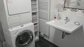 A compact laundry area with a front-loading washing machine, a drying rack, and a sink with a mirror above it.