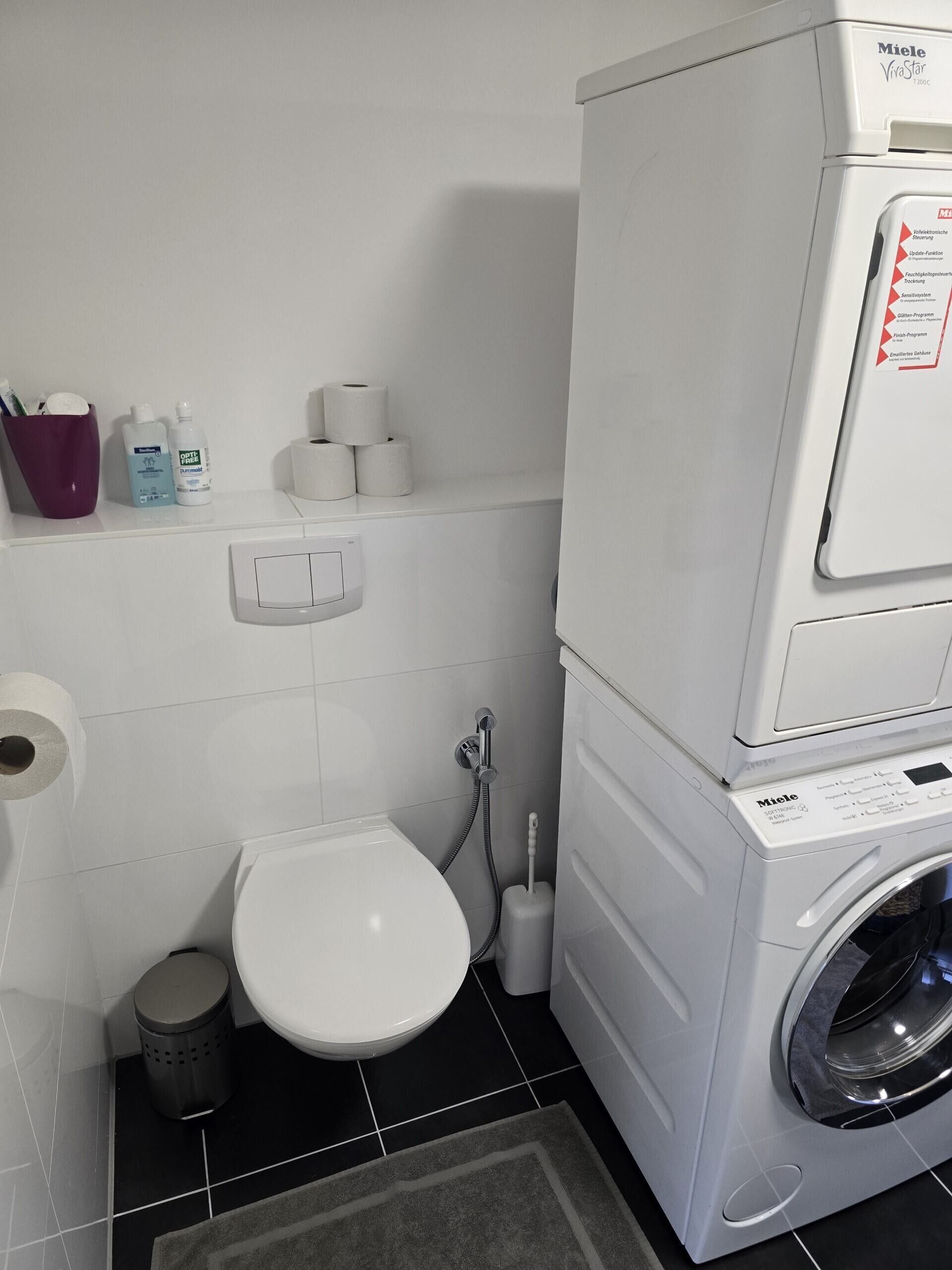 A compact laundry area with a washing machine, a toilet, and a small trash bin.