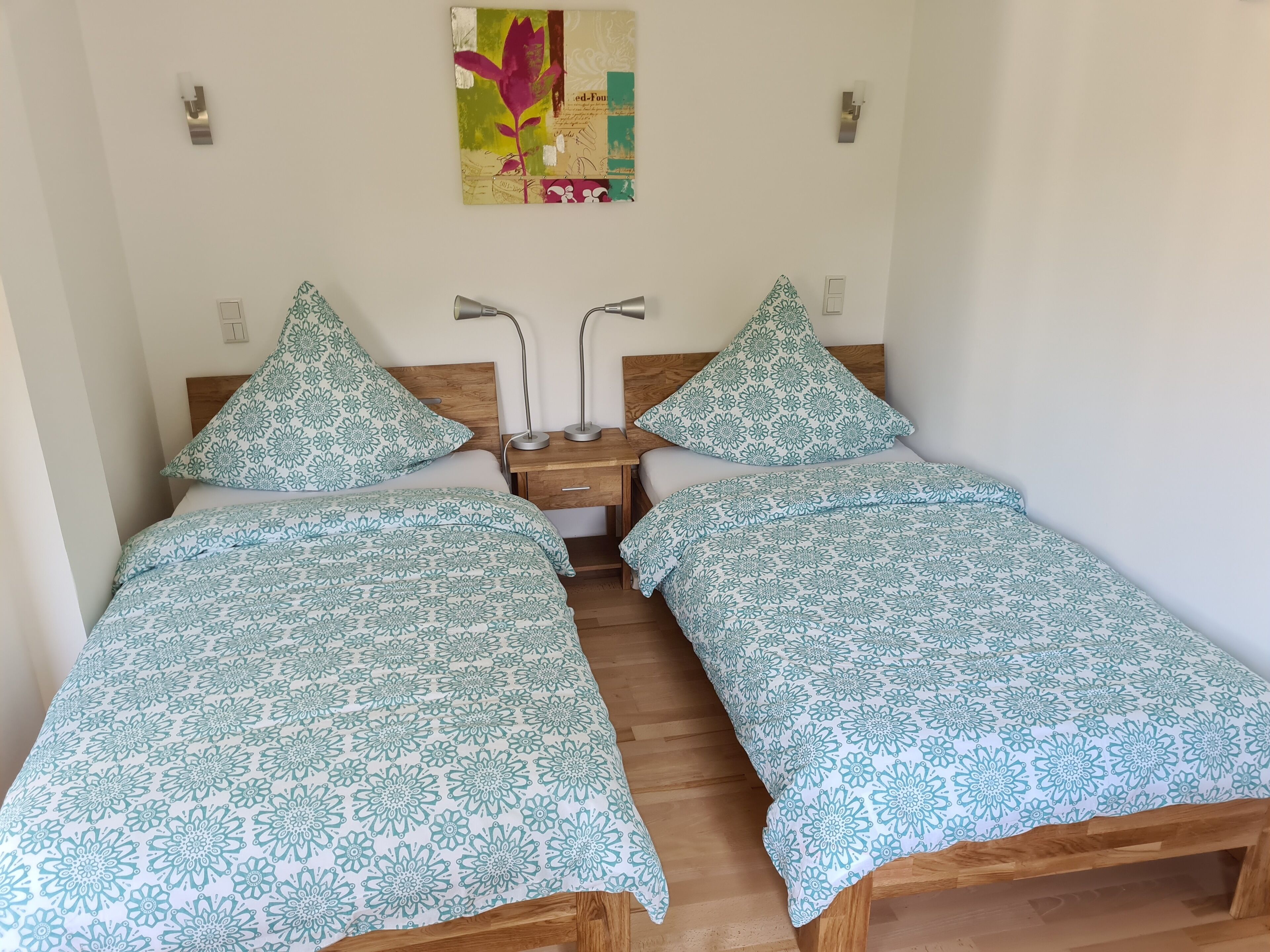 A room with two beds, each with a floral-patterned bedspread, a wooden headboard, a nightstand with a lamp, and a wall-mounted artwork.