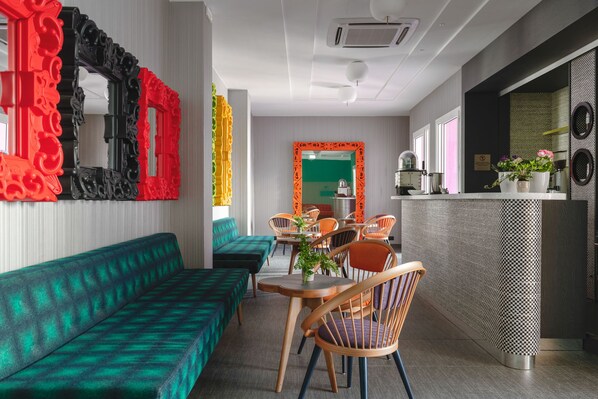 Un hall d’hôtel moderne avec un canapé vert, des chaises en bois, un mur à carreaux et un coin cuisine visible à travers une porte ouverte.