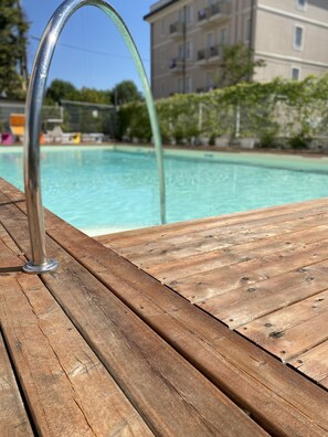 A wooden deck with a metal poolside chair and a pool in the background. - Hotel Rainbow (Rimini)