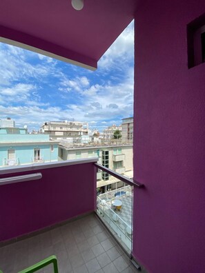 Un balcon avec un mur violet, une rambarde en verre et une vue sur d’autres immeubles et une piscine bleue.