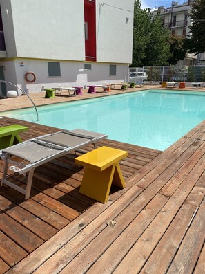 Un espace piscine avec une terrasse en bois, un tabouret jaune et une chaise longue en métal.