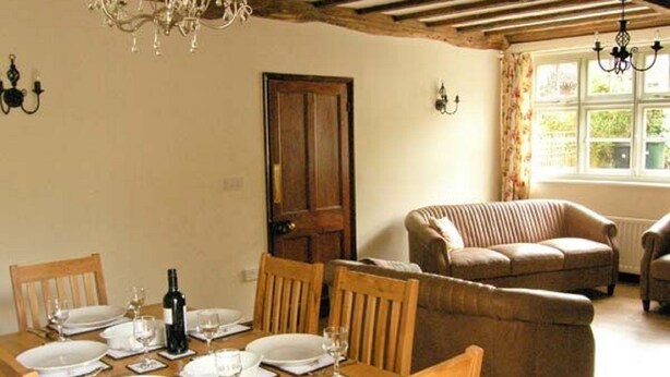 Un comedor con una mesa de madera dispuesta para seis personas, rodeada de sillas de madera. Se observa un sofá marrón y una ventana con cortinas al fondo.