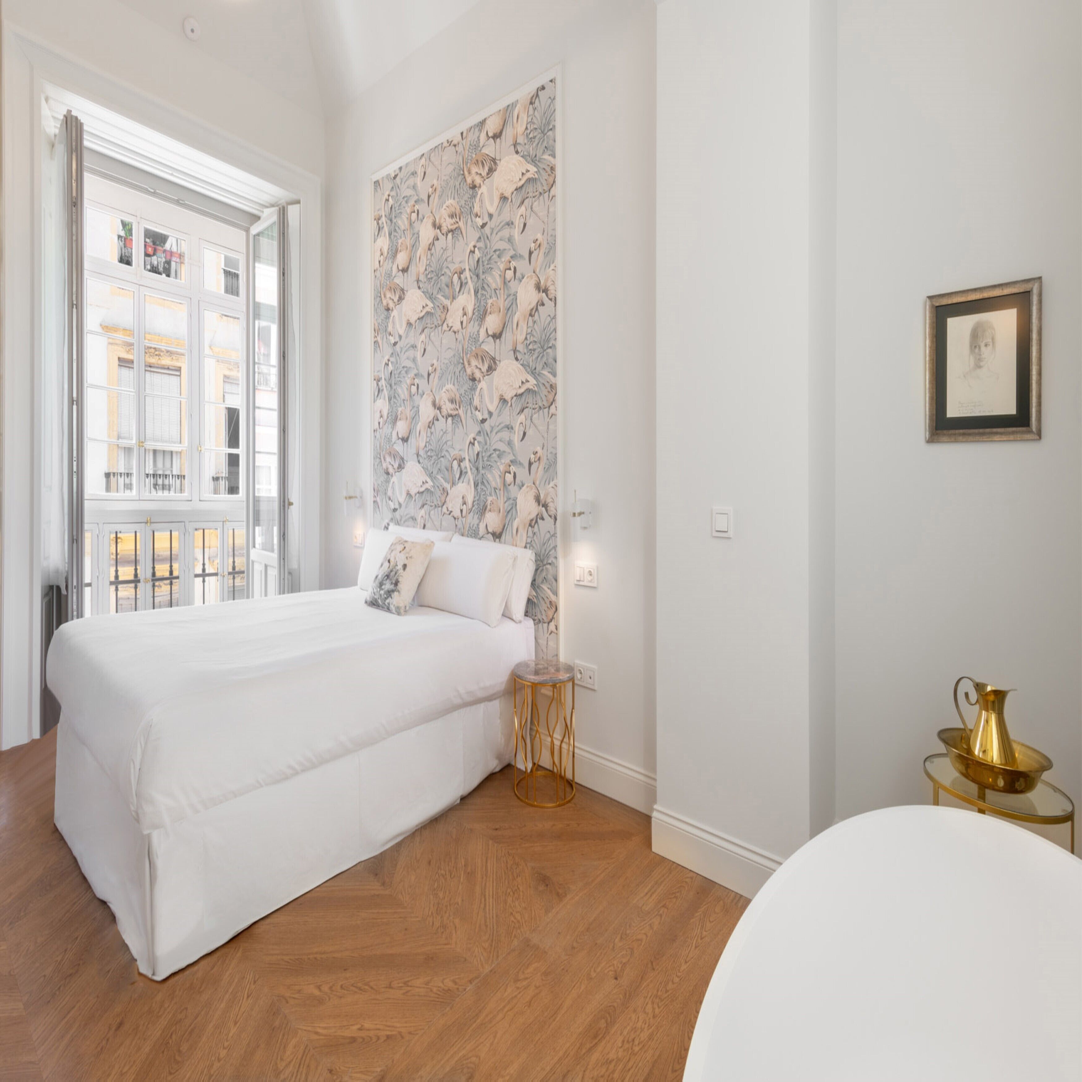 A bedroom with a large bed, a window, a wall with a floral pattern, a small round table with a gold pitcher, and a framed picture on the wall.