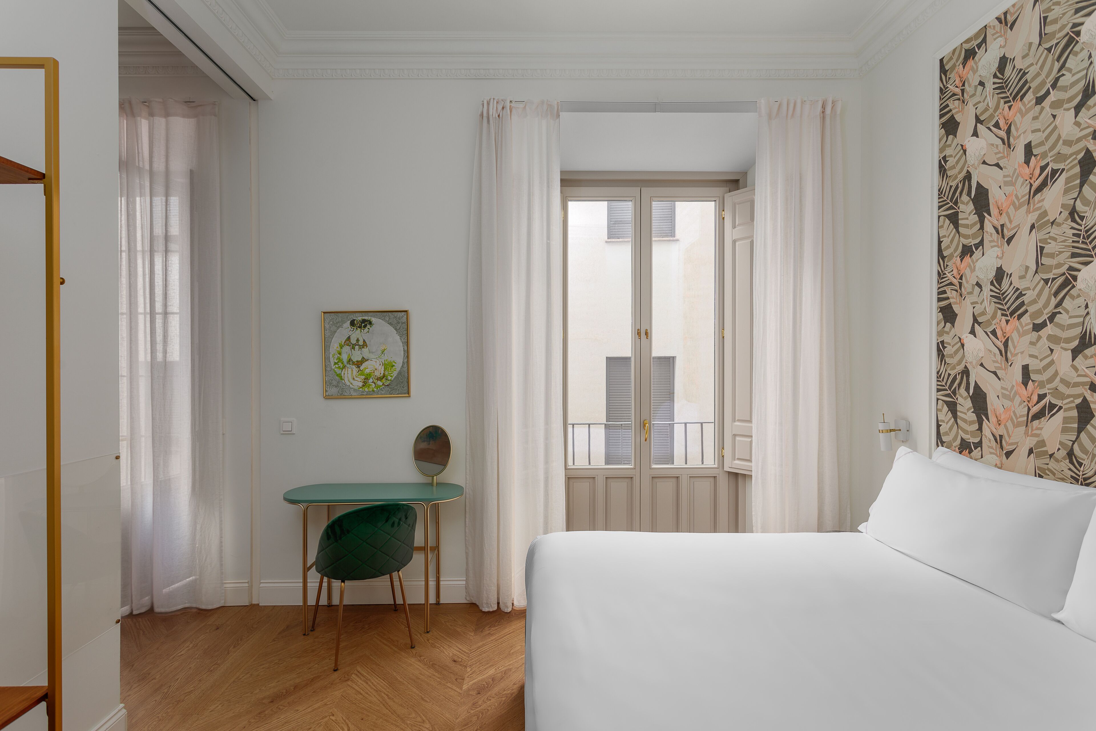 Habitación de hotel con cama, escritorio con silla, ventana con cortinas y pared con estampado de hojas.