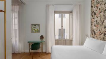A hotel room with a bed, a desk with a chair, a window with curtains, and a wall with a leaf pattern.