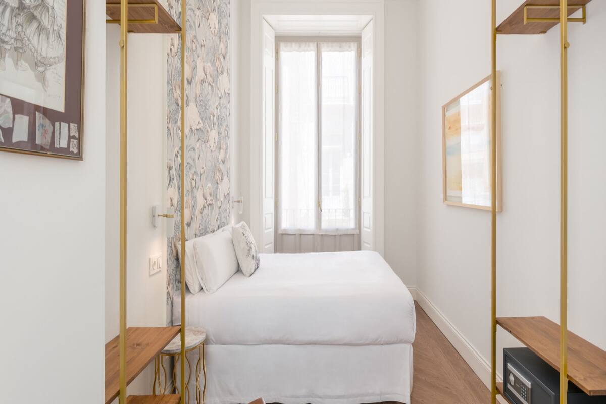 Une chambre moderne avec un lit, un bureau et une chaise. On y trouve une fenêtre avec des rideaux, un mur orné d’un motif floral et un tableau encadré accroché au mur.