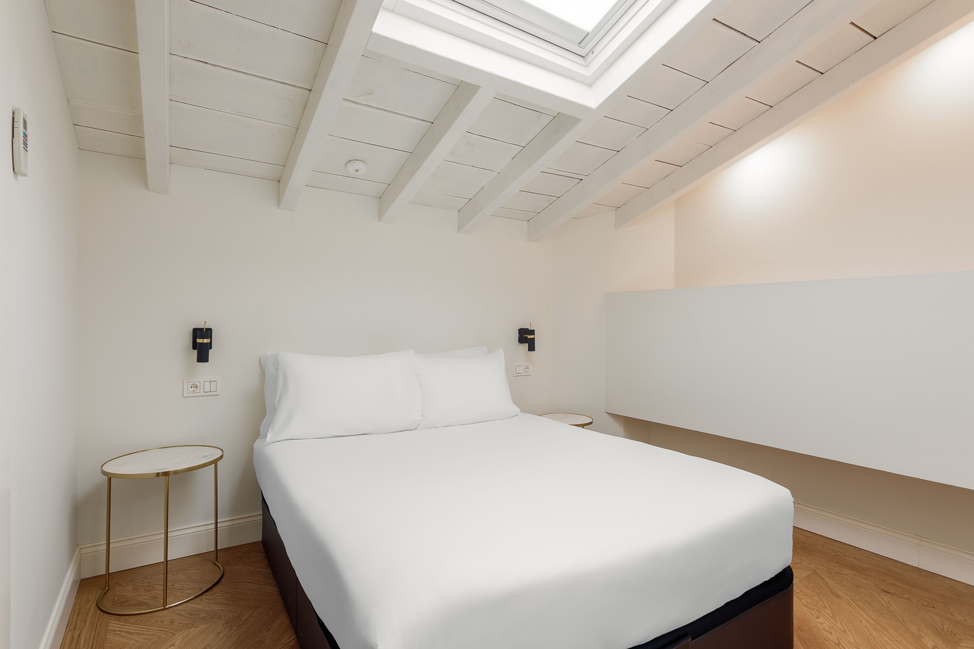 Une chambre à coucher avec un lit, une petite table ronde, une lampe fixée au mur et un puits de lumière.