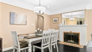 A dining room with a fireplace, white chairs, a chandelier, and a painting on the wall.