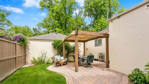 A backyard patio with a wooden pergola, a dining table with chairs, and a small seating area with a sofa and a side table.