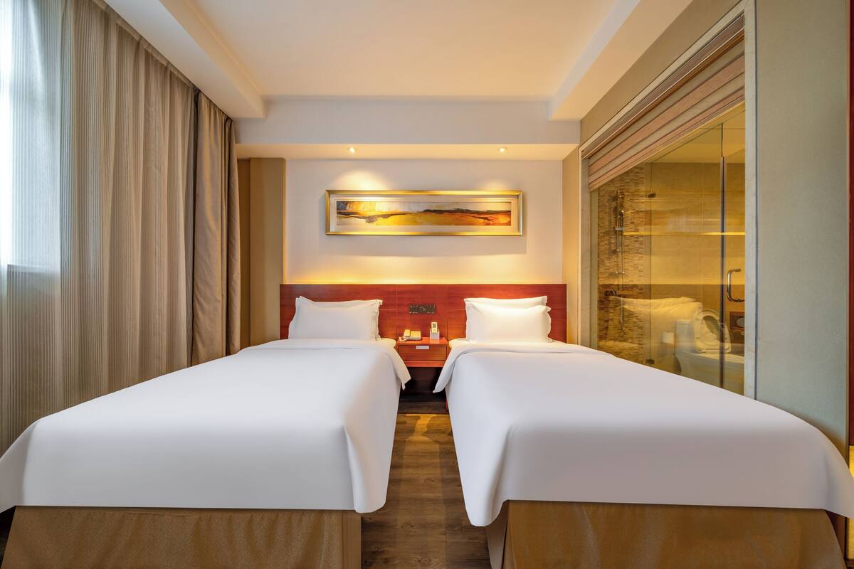 A hotel room with two beds, a painting on the wall, and a glass shower enclosure.