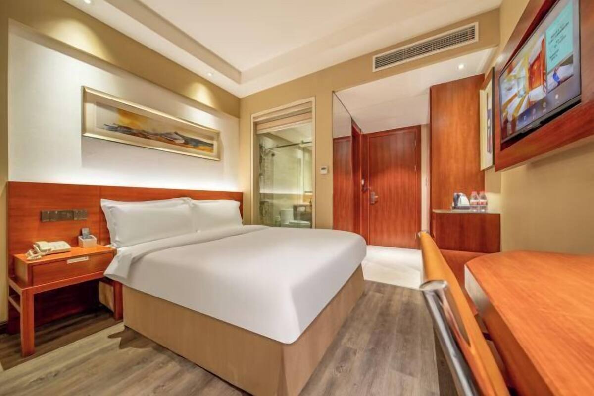 A hotel room with a bed, bedside table, a mirror, a television, and a wooden wardrobe.