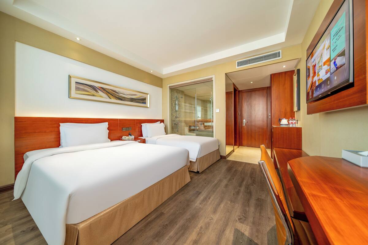 A hotel room with two beds, a wooden headboard, a TV, a desk, and a bathroom visible through a glass door.