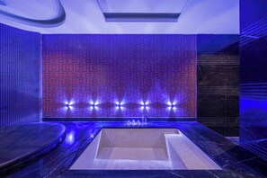 Een moderne spa met een ruime badkuip, een zithoek en een muur met een blauw verlicht mozaïekpatroon.