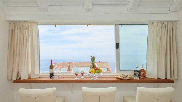 A room with a large window overlooking a cityscape and the sea. There is a wooden table with a bottle of wine, glasses, and a fruit bowl. Three white chairs are positioned in front of the table.